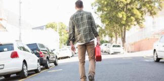 Ein Mann geht zu Fuß eine asphaltierte Straße entlang und trägt in seiner rechten Hand einen roten Benzinkanister. Er macht sich auf den Weg von der Tankstelle zu seinem Auto, um es wieder vollzutanken.