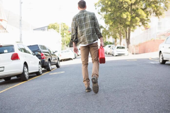 Ein Mann geht zu Fuß eine asphaltierte Straße entlang und trägt in seiner rechten Hand einen roten Benzinkanister. Er macht sich auf den Weg von der Tankstelle zu seinem Auto, um es wieder vollzutanken.