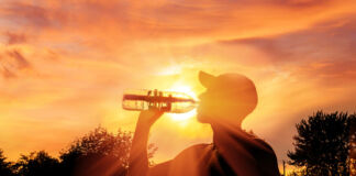 Ein Mann mit Cappy trinkt in den Strahlen eines wunderschönen abendlichen Sonnenuntergangs am Waldrand und einem Sportplatz eine Flasche Wasser. Die Sonnenstrahlen durchscheinen die Flasche und strahlen hell in die Kamera.