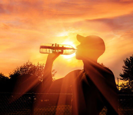 Ein Mann mit Cappy trinkt in den Strahlen eines wunderschönen abendlichen Sonnenuntergangs am Waldrand und einem Sportplatz eine Flasche Wasser. Die Sonnenstrahlen durchscheinen die Flasche und strahlen hell in die Kamera.