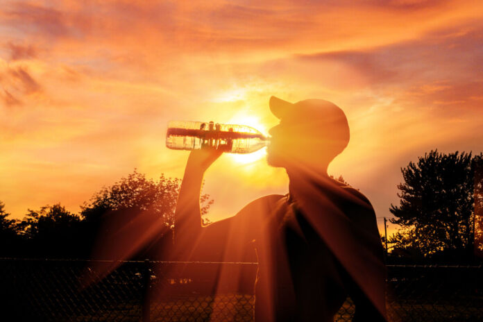 Ein Mann mit Cappy trinkt in den Strahlen eines wunderschönen abendlichen Sonnenuntergangs am Waldrand und einem Sportplatz eine Flasche Wasser. Die Sonnenstrahlen durchscheinen die Flasche und strahlen hell in die Kamera.