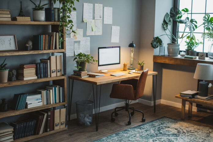 Ein modern eingerichtetes Büro, in dem alles vorzufinden ist, was man zum Arbeiten im Home-Office braucht. Ein Schreibtischstuhl, ein Bildschirm, ein Bücherregal mit Aktenordner, Zimmerpflanzen und weiteren Dekoartikeln und Notizen an der Wand. Ein schöner Teppich rundet die Optik des Büros ab.