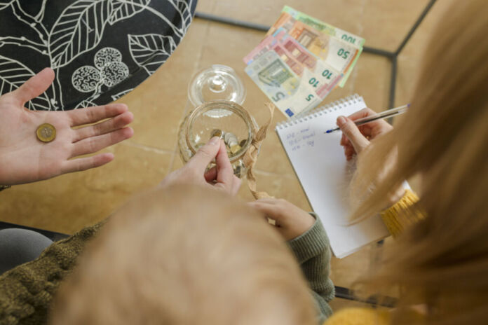 Man sieht aus der Vogelperspektive, dass mehrere Familienmitglieder verschiedenen Alters am Tisch sitzen. Vor ihnen liegen einige wenige Euro-Scheine und ein Notizheft. Die Erwachsenen rechnen ihre Ausgaben und Einnahmen gegeneinander und erstellen einen Finanzplan.