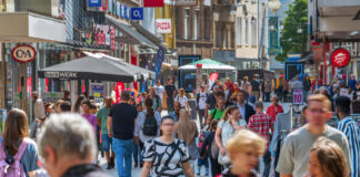 Eine belebte Fußgängerzone an einem warmen Sommertag. Die Straße ist voller Menschen und rechts und links sieht man mehrere Geschäfte, in denen man einkaufen kann. Zudem befinden sich viele Cafés und Restaurants entlang des Fußgängerwegs.