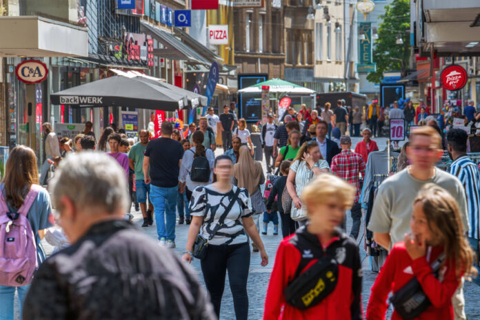 Eine belebte Fußgängerzone an einem warmen Sommertag. Die Straße ist voller Menschen und rechts und links sieht man mehrere Geschäfte, in denen man einkaufen kann. Zudem befinden sich viele Cafés und Restaurants entlang des Fußgängerwegs.