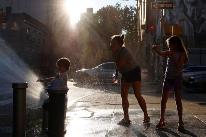 Eine erwachsene Frau und zwei Kinder stehen an einem Wasserhydranten und gönnen sich eine Abkühlung. Es scheint sehr warm zu sein und die Sonne blendet und taucht alles in ein helles Licht.