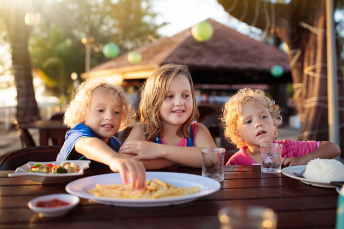 Drei Kinder essen etwas gemeinsam, sie haben Spaß miteinander. Es handelt sich wohl um eine kinderfreundliche Mahlzeit, denn allen scheint es zu schmecken.