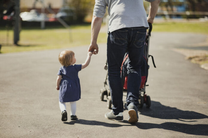 Ein Vater hält seine kleine Tochter an der Hand und geht mit ihr wohl in einem Park spazieren. Mit der anderen Hand schiebt er den Kinderwagen.