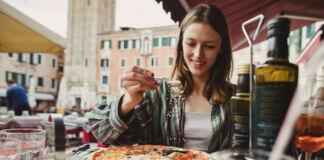 Eine Frau sitzt in der Innenstadt in einem Restaurant und hat einen Teller mit einer großen Pizza vor sich stehen. Die Frau streut mit einem Löffel Käse auf ihr Essen,