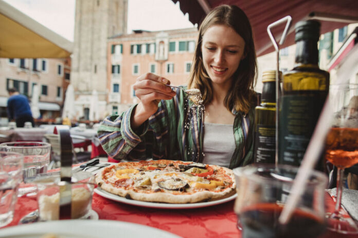 Eine Frau sitzt in der Innenstadt in einem Restaurant und hat einen Teller mit einer großen Pizza vor sich stehen. Die Frau streut mit einem Löffel Käse auf ihr Essen,