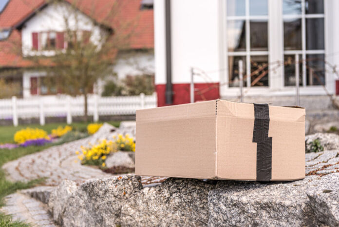 Ein Paket aus Karton liegt in einem Garten auf einem Gehweg. Das Paket ist mit Paketklebeband verschlossen. Im Hintergrund kann man Blumen und einen weißen Zaun sowie das Nachbarhaus erkennen.