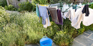 Frisch gewaschene Wäsche – Handtücher und Kleidung – hängt auf einer Wäschespinne an einem sonnigen Tag im Hintergarten. Darunter steht ein blauer Wäschekorb. Die Wäsche trocknet rasch im Sonnenschein.