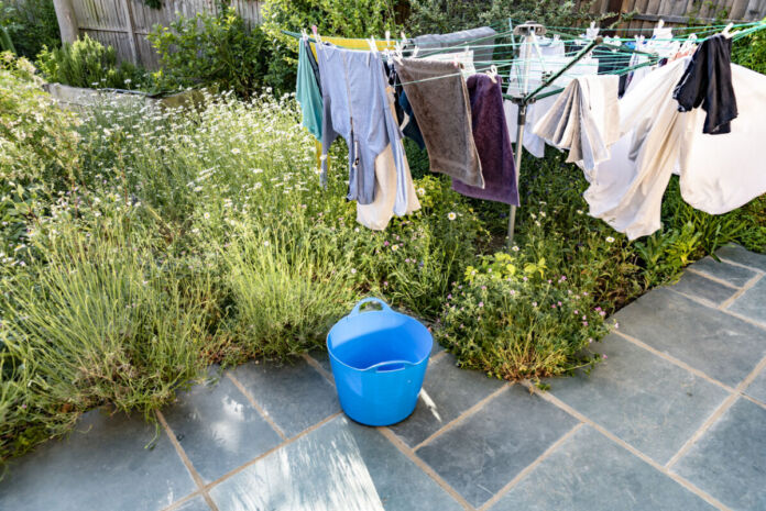 Frisch gewaschene Wäsche – Handtücher und Kleidung – hängt auf einer Wäschespinne an einem sonnigen Tag im Hintergarten. Darunter steht ein blauer Wäschekorb. Die Wäsche trocknet rasch im Sonnenschein.