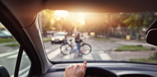 Das Bild zeigt die Perspektive des Autofahrers am Steuer. Eine Hand mit Armbanduhr liegt auf dem Lenkrad. Vor dem Auto ist verschwommen eine Fahrradfahrerin, die ihr Rad über einen Zebrastreifen schiebt.