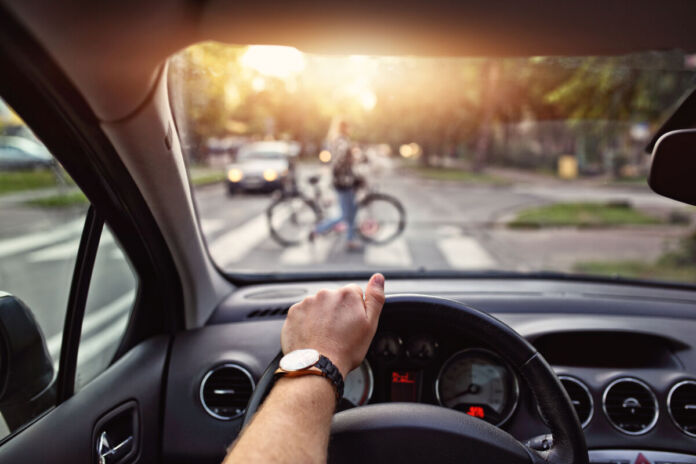 Das Bild zeigt die Perspektive des Autofahrers am Steuer. Eine Hand mit Armbanduhr liegt auf dem Lenkrad. Vor dem Auto ist verschwommen eine Fahrradfahrerin, die ihr Rad über einen Zebrastreifen schiebt.