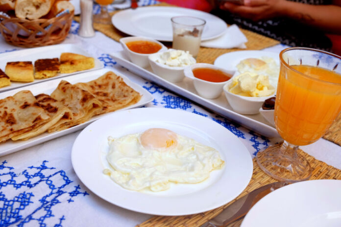 Auf einem gedeckten Tisch stehen verschiedene Speisen zum Frühstück. Im Vordergrund ein gebratenes Ei auf weißem Teller, daneben Börek oder Blätterteig, weitere Gebäcke, Marmelade, Butter, Quark sowie ein Glas mit Multivitaminsaft. Die Atmosphäre wirkt sehr sommerlich.