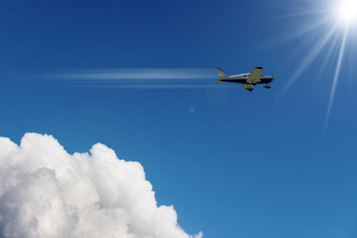 Ein kleines schwarzes Flugzeug fliegt über den Wolken. Es zieht einen weißen Kondensstreifen hinter sich her. Die Sonne scheint vom Himmel.