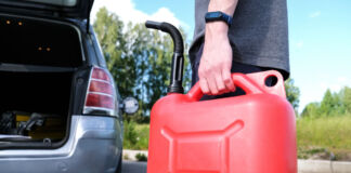 Ein Mann hält in der linken Hand einen roten Gas-Kanister, mit dem er gerade zu seinem Auto läuft, um es zu betanken. Im Hintergrund sieht man das Auto mit geöffnetem Kofferraum und geöffneten Türen.