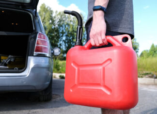 Ein Mann hält in der linken Hand einen roten Gas-Kanister, mit dem er gerade zu seinem Auto läuft, um es zu betanken. Im Hintergrund sieht man das Auto mit geöffnetem Kofferraum und geöffneten Türen.