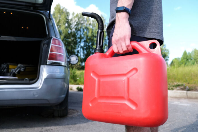 Ein Mann hält in der linken Hand einen roten Gas-Kanister, mit dem er gerade zu seinem Auto läuft, um es zu betanken. Im Hintergrund sieht man das Auto mit geöffnetem Kofferraum und geöffneten Türen.