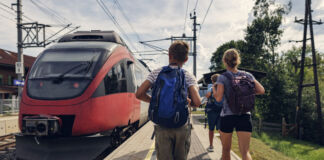 Eine Familie läuft an einem Bahnsteig entlang. Die Frau und der Junge tragen Rucksäcke, da sie in den Urlaub fahren. Neben ihnen steht ein roter Zug am Bahngleis des Bahnhofs.