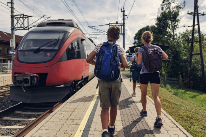 Eine Familie läuft an einem Bahnsteig entlang. Die Frau und der Junge tragen Rucksäcke, da sie in den Urlaub fahren. Neben ihnen steht ein roter Zug am Bahngleis des Bahnhofs.