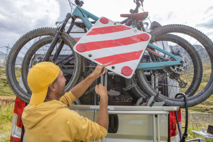 Ein Mann befestigt ein Fahrrad auf einem Fahrradträger, um Fährräder mit dem Auto zu transportieren. Er bringt auch ein Warnzeichen an.