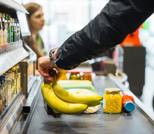 Eine Person geht im Supermarkt einkaufen. Sie legt Bananen und weitere Artikel auf das Band an der Kasse. Der Kunde bezahlt und verlässt den Supermarkt.