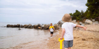 Zwei kleine Jungen spielen an einem Strand am Meer. Sie rennen durch den Sand, in dem sich Muscheln befinden. Einer der Jungen zieht einen Spielzeugwagen mit Sandspielzeug hinter sich her.