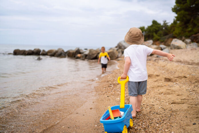 Zwei kleine Jungen spielen an einem Strand am Meer. Sie rennen durch den Sand, in dem sich Muscheln befinden. Einer der Jungen zieht einen Spielzeugwagen mit Sandspielzeug hinter sich her.