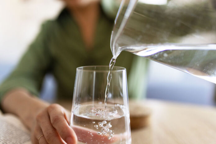 Eine Frau gießt in ein Glas Wasser aus einer durchsichtigen Karaffe ein. Während das Glas, die Hand sowie die Karaffe deutlich zu erkennen sind, befindet sich die Frau im Hintergrund ist verschwommen.