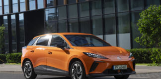 Ein modern geschnittenes orange -farbiges E-Auto der Marke MG steht auf der Straße vor einem modernen Gebäude aus Stahl und Glas.