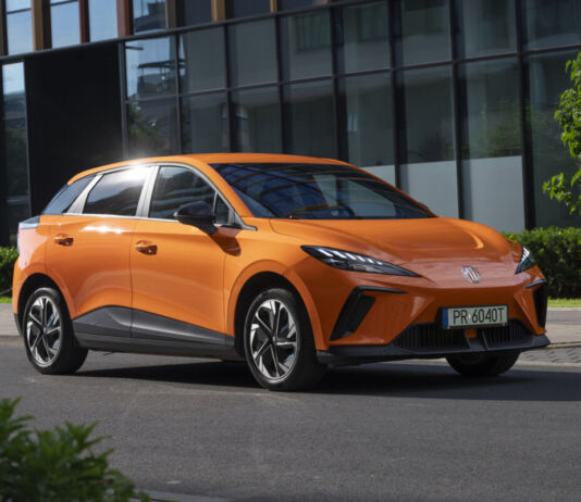 Ein modern geschnittenes orange -farbiges E-Auto der Marke MG steht auf der Straße vor einem modernen Gebäude aus Stahl und Glas.