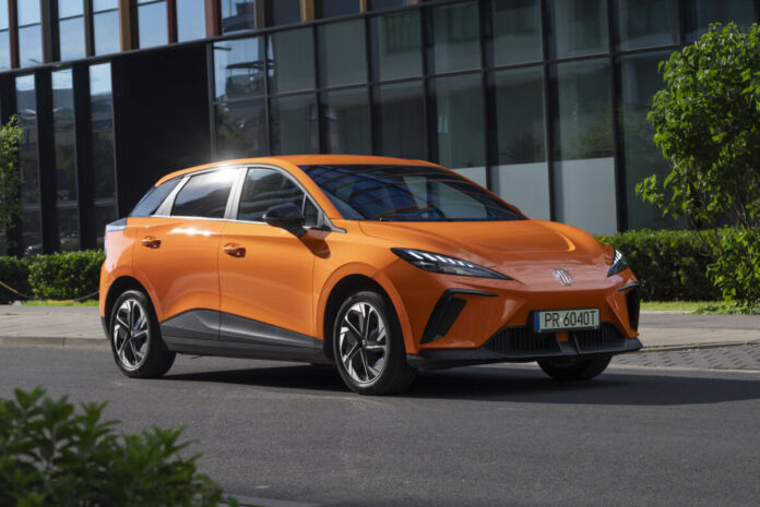 Ein modern geschnittenes orange -farbiges E-Auto der Marke MG steht auf der Straße vor einem modernen Gebäude aus Stahl und Glas.