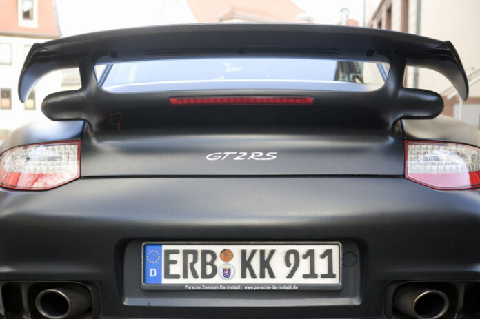 Man blickt von hinten auf einen schwarzes Auto von Porsche, sodass man den Auspuff, die Rücklichter, einen Spoiler sowie das Nummernschild erkennen kann. Das Autokennzeichen ist aus Deutschland.