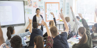 In einem Klassenraum sitzen die Schülerinnen und Schüler auf ihren Plätzen und schauen nach vorn. Viele von ihnen melden sich. Vor der Klasse steht eine Lehrerin und lächelt die Klasse an.