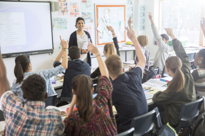 In einem Klassenraum sitzen die Schülerinnen und Schüler auf ihren Plätzen und schauen nach vorn. Viele von ihnen melden sich. Vor der Klasse steht eine Lehrerin und lächelt die Klasse an.