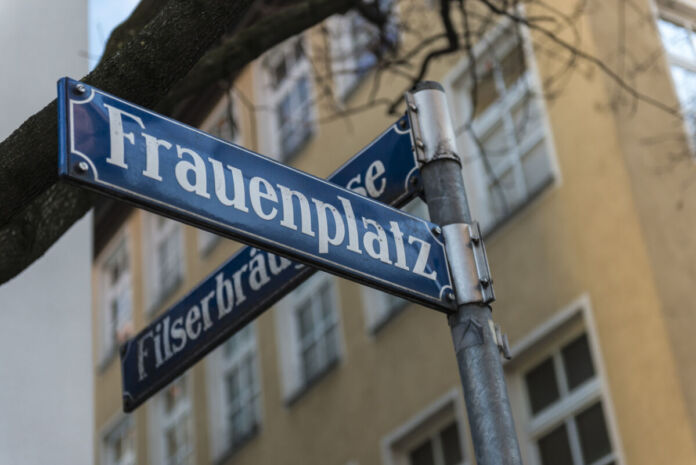 Vor einem Haus mit gelbem Anstrich steht ein Straßenschild mit zwei Straßennamen. Auf dem blauen Schild kann man die Bezeichnungen von zwei Straßen lesen.