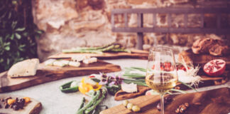 Ein reich gedeckter Tisch mit Wein, Obst und Gemüse lädt zum Verweilen und Essen ein. Im Hintergrund liegt frisches Brot. Das sieht nach einem Picknick im Sommer aus.