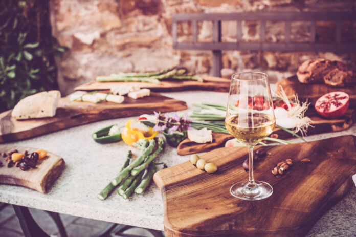 Ein reich gedeckter Tisch mit Wein, Obst und Gemüse lädt zum Verweilen und Essen ein. Im Hintergrund liegt frisches Brot. Das sieht nach einem Picknick im Sommer aus.