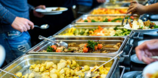In mehreren großen Stahlschüsseln sind verschiedene Gerichte wie buntes Gemüse und Kartoffeln angerichtet. Die Menschen können sich in der Kantine an den Mahlzeiten bedienen.