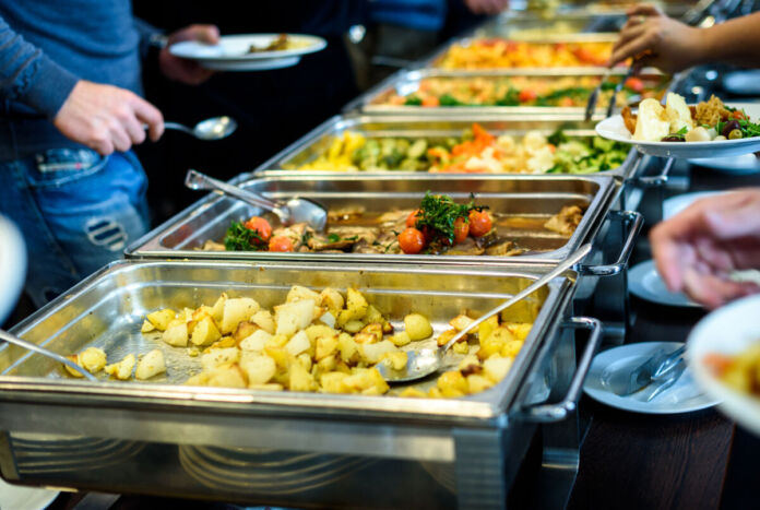 In mehreren großen Stahlschüsseln sind verschiedene Gerichte wie buntes Gemüse und Kartoffeln angerichtet. Die Menschen können sich in der Kantine an den Mahlzeiten bedienen.