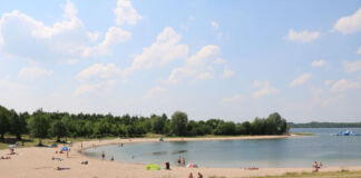 Der Schladitzer See nahe Leipzig ist ein beliebter Urlaubsort. Mehrere Menschen befinden sich an einem Badestrand in Bikini und Badehose. Manche schwimmen im Wasser. Manche sonnen sich auf Badetüchern.