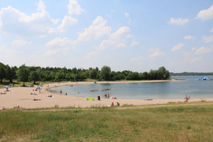 Der Schladitzer See nahe Leipzig ist ein beliebter Urlaubsort. Mehrere Menschen befinden sich an einem Badestrand in Bikini und Badehose. Manche schwimmen im Wasser. Manche sonnen sich auf Badetüchern.