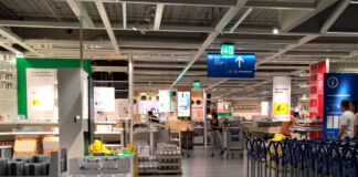 Ein Blick in eine Filiale von dem schwedischen Möbelhaus IKEA. Man kann die Einkaufswagen erkennen sowie einige Kunden, die gerade dabei sind, einzukaufen. Es stapeln sich Paletten mit Wohnzubehör und Küchenutensilien direkt am Eingang zu dem Einkaufsbereich.