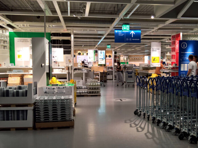 Ein Blick in eine Filiale von dem schwedischen Möbelhaus IKEA. Man kann die Einkaufswagen erkennen sowie einige Kunden, die gerade dabei sind, einzukaufen. Es stapeln sich Paletten mit Wohnzubehör und Küchenutensilien direkt am Eingang zu dem Einkaufsbereich.