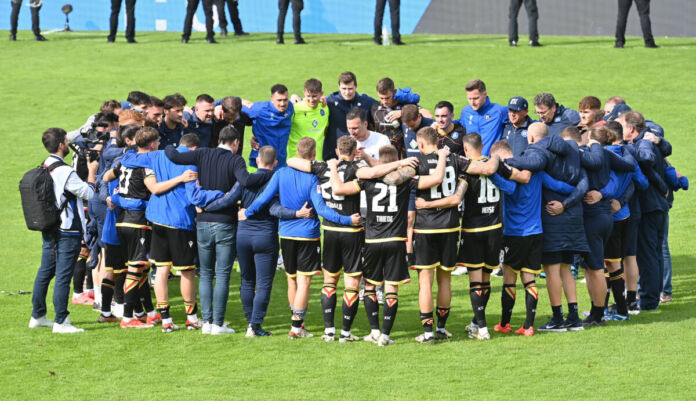 Mehrere Spieler einer Fußballmannschaft stehen gemeinsam Arm in Arm in ihren Trikots auf dem Fußballplatz. Die Bundesliga-Spieler bereiten sich auf das Fußballspiel vor.