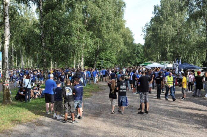 Viele Karlsruher SC Fans strömen zum Fußballspiel ihres Vereins. Sie tragen Fan-Shirts und Trikots. Die Kleidung und die Fanartikel sind in den Vereinsfarben blau und weiß gehalten.