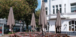 Ein Restaurant oder eine Gaststätte scheint geschlossen zu haben. In der Tür hängt ein Zettel, dessen Aufschrift man nicht lesen kann. Die Sitzbänke und Tische aus Holz sind alle dicht zusammengeschoben und mit rot-weißem Absperrband versehen. Die Sonnenschirme sind zusammengeklappt und ebenfalls mit dem Band versehen.