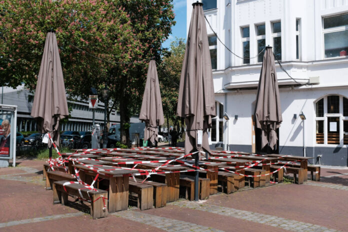 Ein Restaurant oder eine Gaststätte scheint geschlossen zu haben. In der Tür hängt ein Zettel, dessen Aufschrift man nicht lesen kann. Die Sitzbänke und Tische aus Holz sind alle dicht zusammengeschoben und mit rot-weißem Absperrband versehen. Die Sonnenschirme sind zusammengeklappt und ebenfalls mit dem Band versehen.